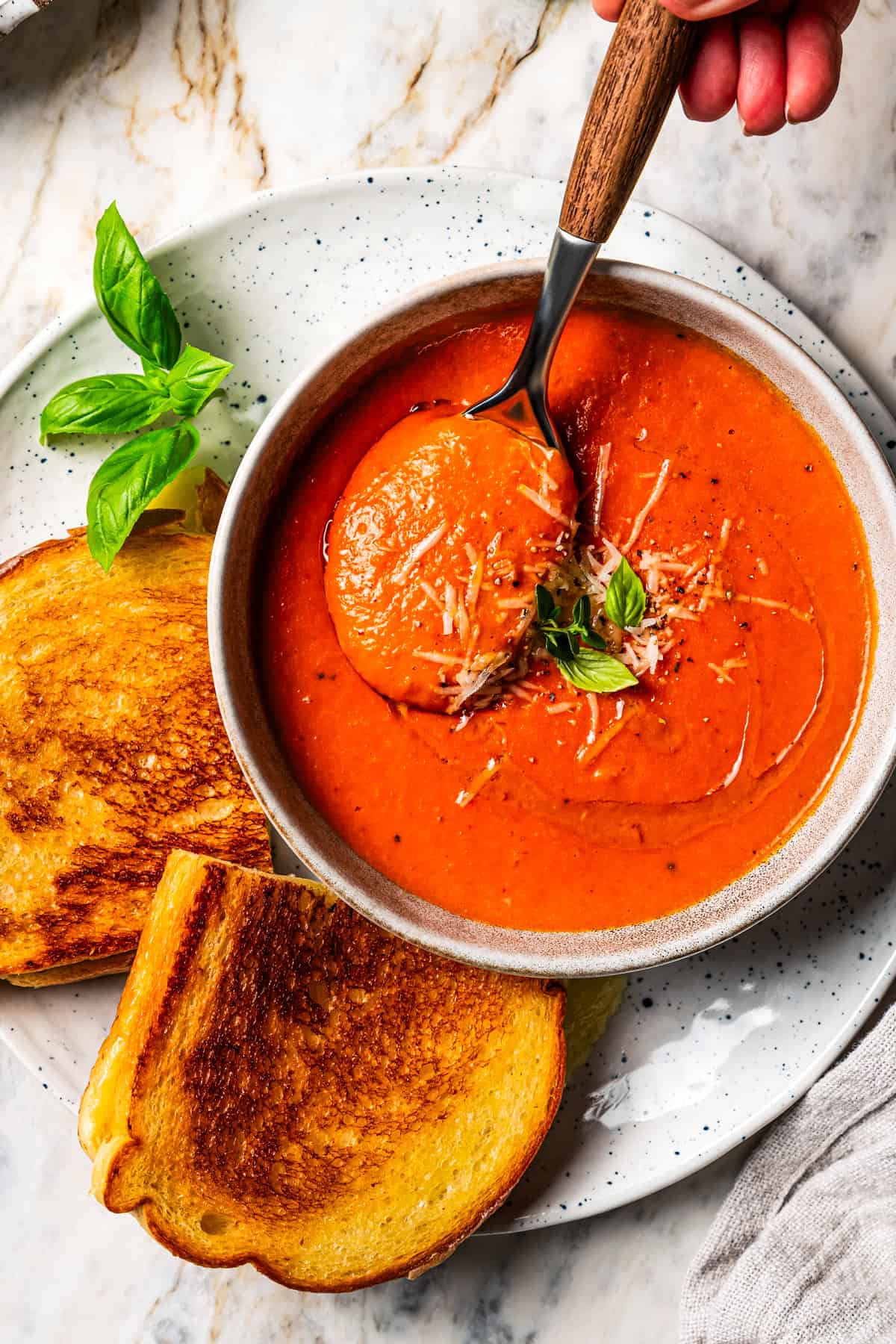 Overhead view of a hand holding a spoon over a bowl of tomato soup, next to a grilled cheese on a plate.