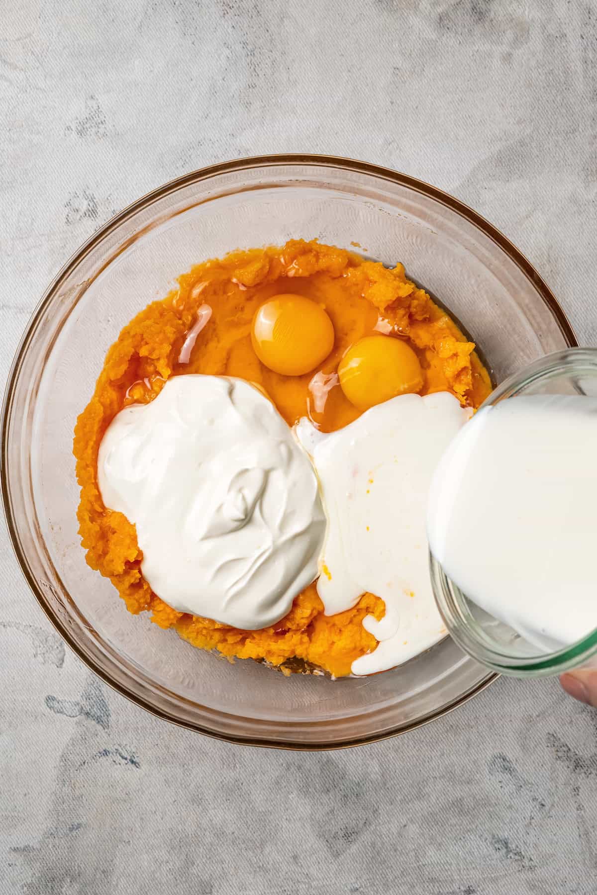 Pouring milk into a bowl of mashed sweet potatoes with eggs and wet ingredients.