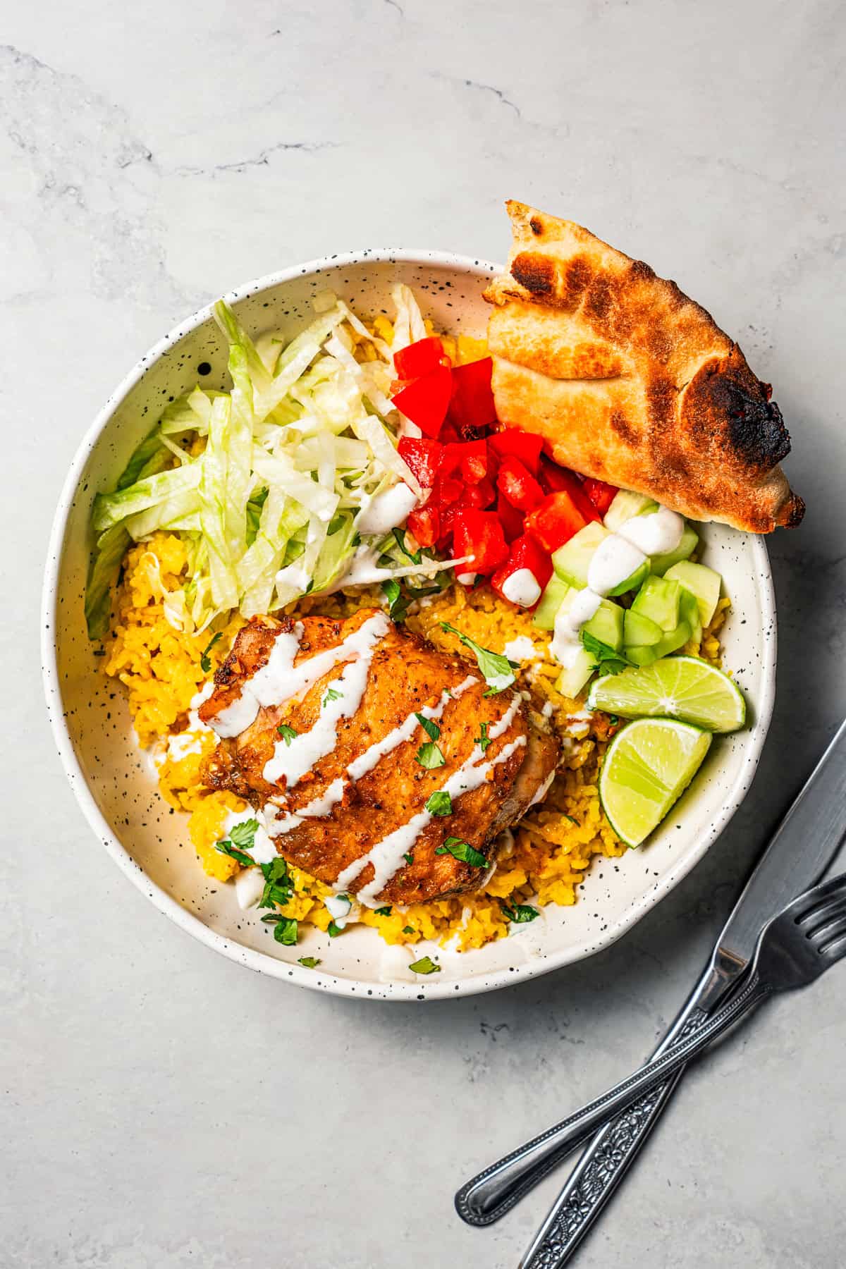 This homemade halal cart chicken over rice combines savory spice-rubbed chicken and fluffy rice, drizzled with the famous creamy white sauce! Overhead view of halal cart chicken over rice in a bowl with flatbread, chopped tomatoes and cucumbers, shredded lettuce, and a sour cream drizzle.
