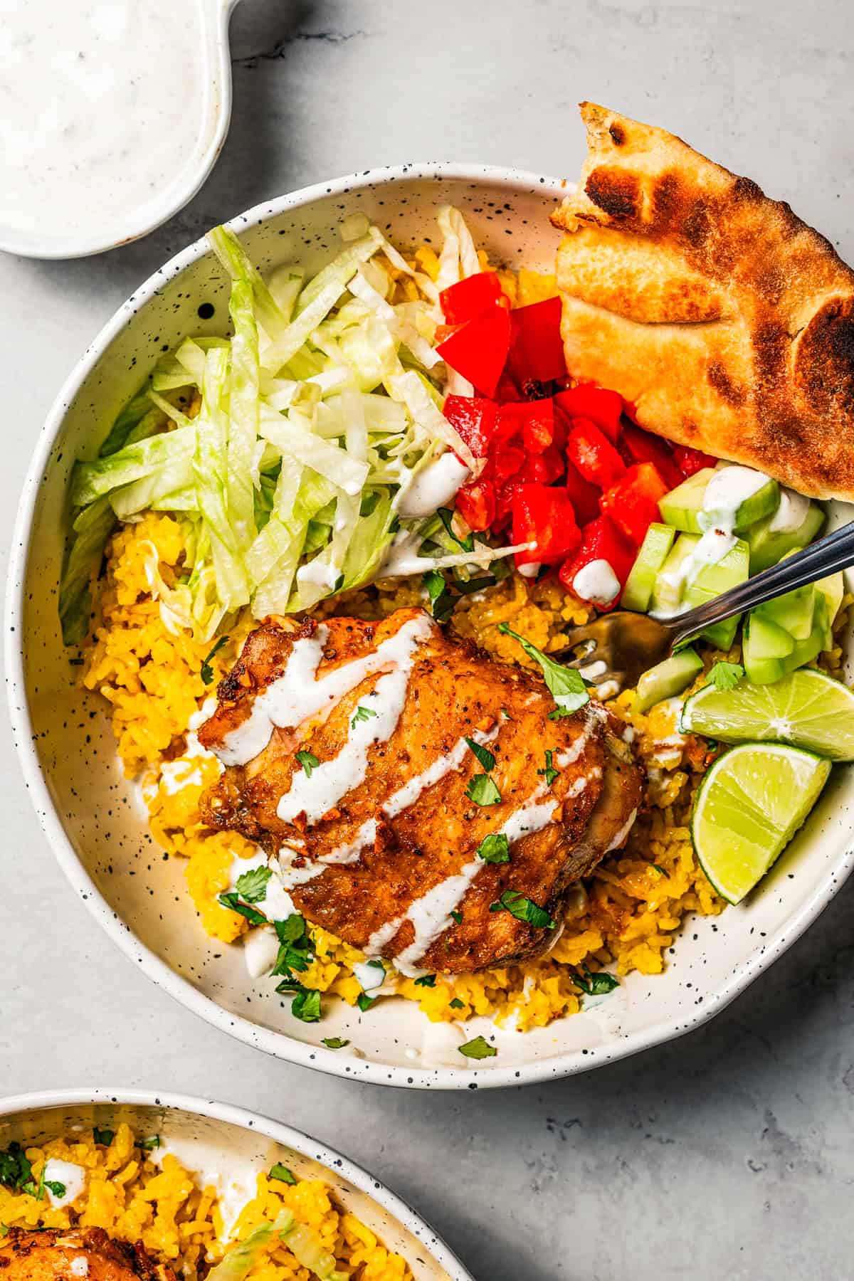 This homemade halal cart chicken over rice combines savory spice-rubbed chicken and fluffy rice, drizzled with the famous creamy white sauce! Halal chicken over rice in a bowl with flatbread, garnishes, and a sour cream drizzle.