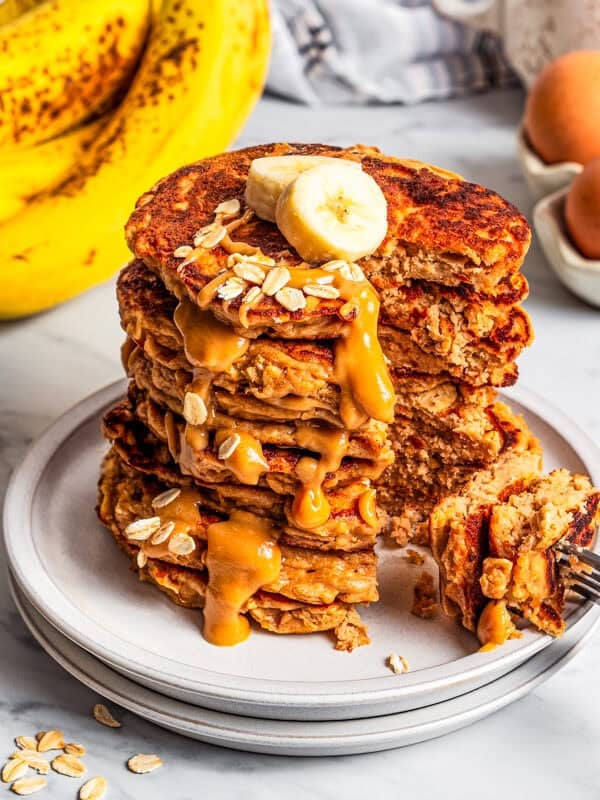 Protein pancakes stacked up on a plate, topped with banana slices and peanut butter maple syrup, with a large bite of pancake stuck on a fork.