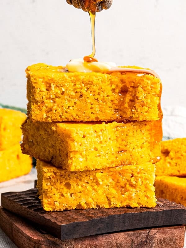 A hand using a honey dipper to drizzle honey over a stack of sweet potato cornbread slices topped with butter, with more slices in the background.
