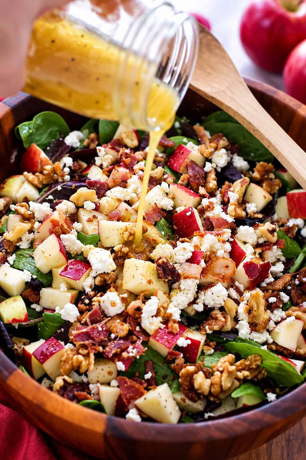 This apple salad recipe combines crisp fall apples tossed with smoky bacon, crunchy walnuts, and creamy feta in a sweet-tangy maple dressing. Maple balsamic dressing being poured over apple bacon salad with mixed greens, feta cheese, and walnuts.