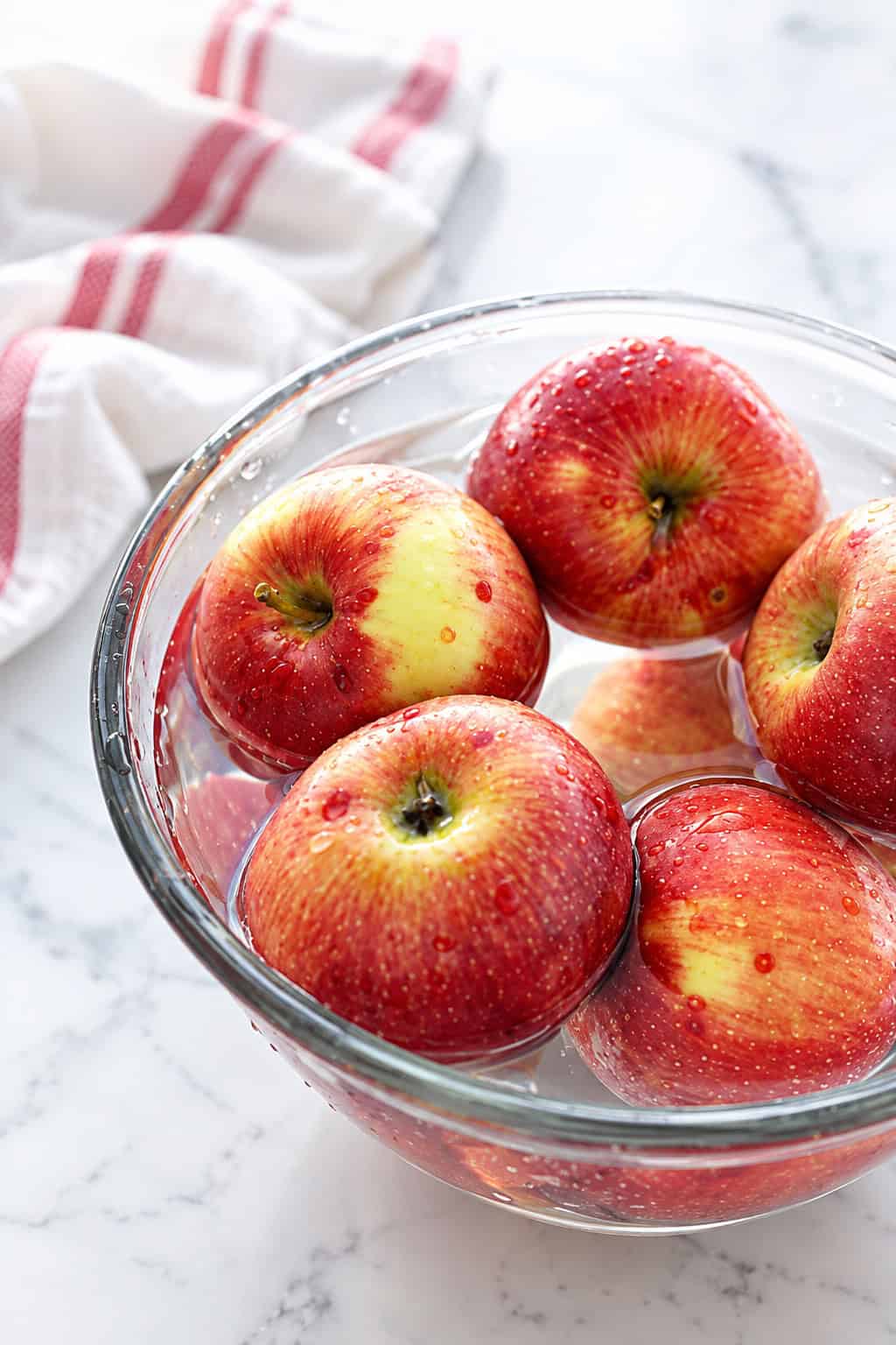 This apple salad recipe combines crisp fall apples tossed with smoky bacon, crunchy walnuts, and creamy feta in a sweet-tangy maple dressing. Red apples soaking in a glass bowl of water on a white marble countertop.