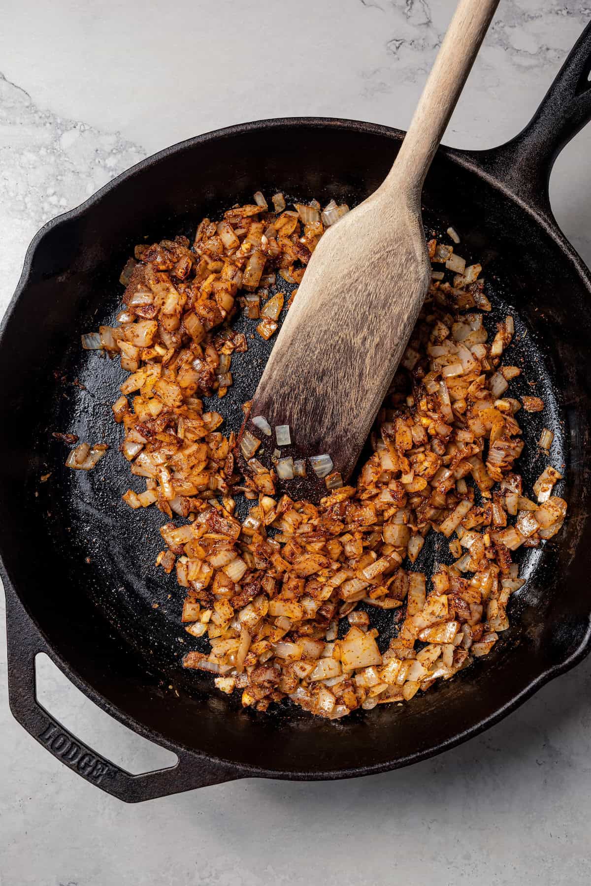 Browned onions with garlic and spices in a skillet with a wooden spatula.