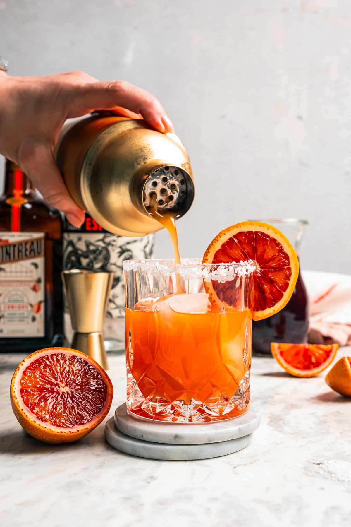 A hand pouring a blood orange margarita from a cocktail shaker into a salt-rimmed glass.