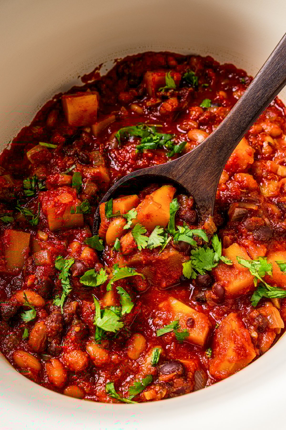 Close-up photo of butternut squash chili garnished with fresh herbs inside the slow cooker with a wooden spoon.
