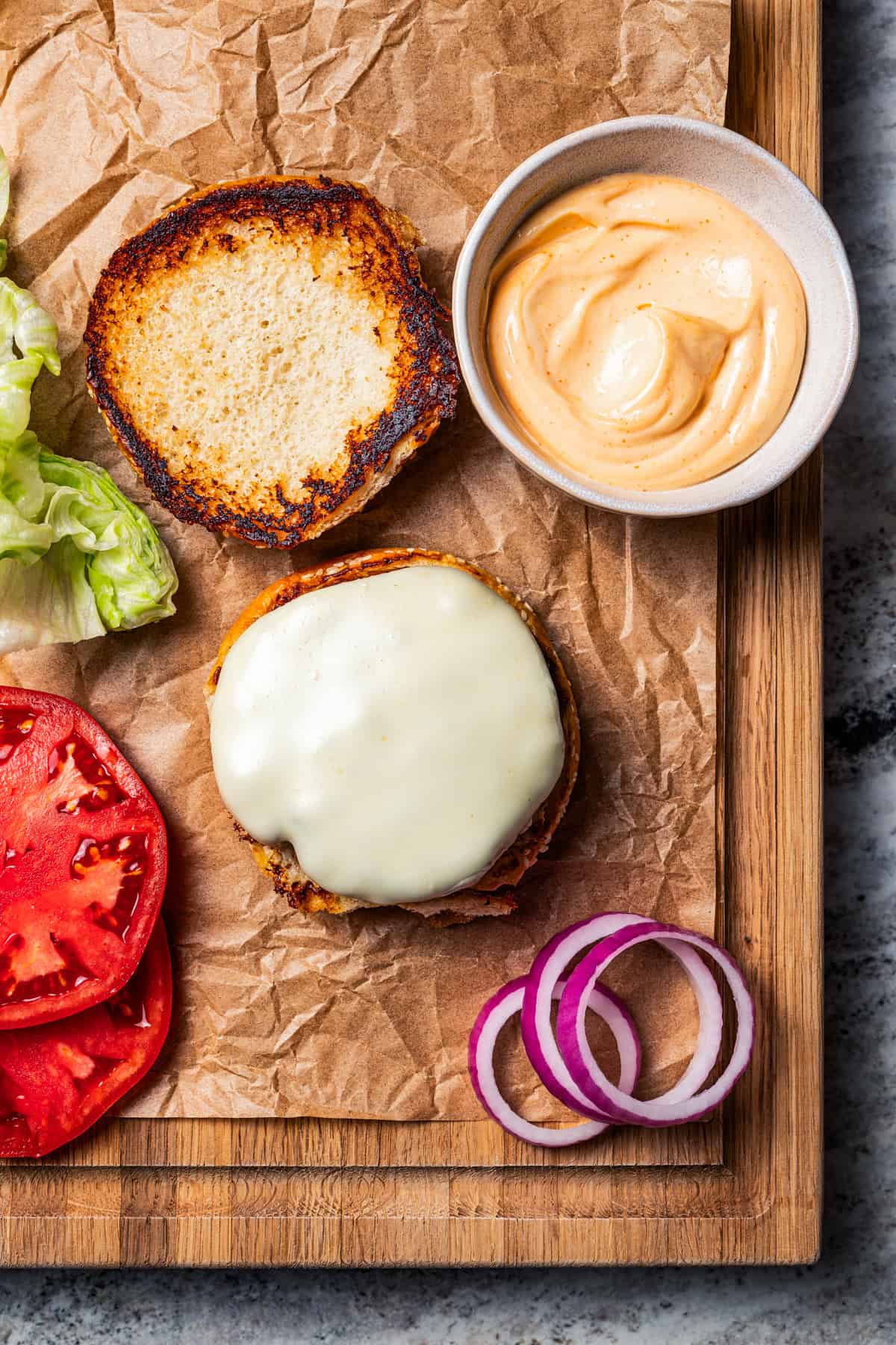 Chicken burger patty topped with melted cheese, surrounded by lettuce, tomatoes, and a bowl of spicy mayo on a wooden cutting board.