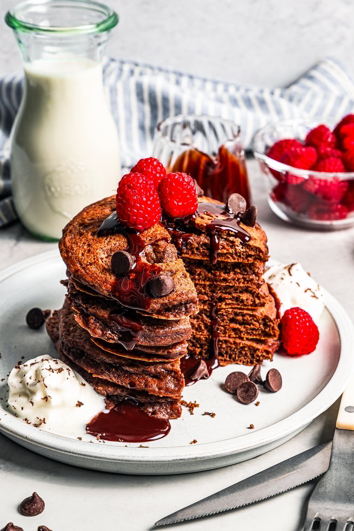 A stack of chocolate pancakes topped with chocolate sauce and fresh raspberries, with a large slice cut out.