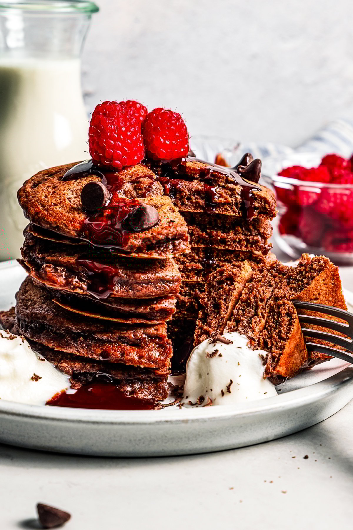 A stack of chocolate pancakes topped with fresh raspberries, with a forkful cut out on a plate.