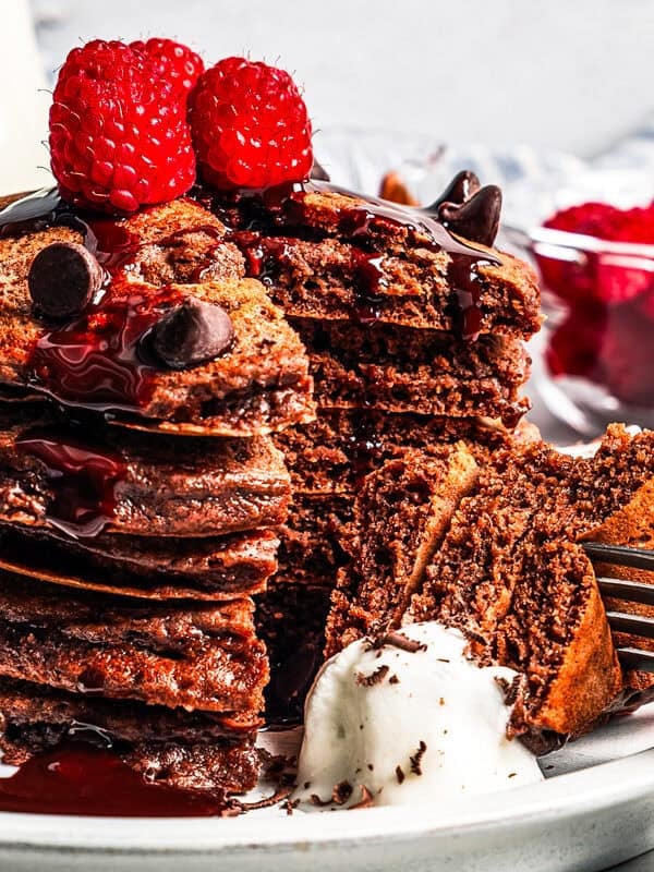 A stack of chocolate pancakes topped with fresh raspberries, with a forkful cut out on a plate.