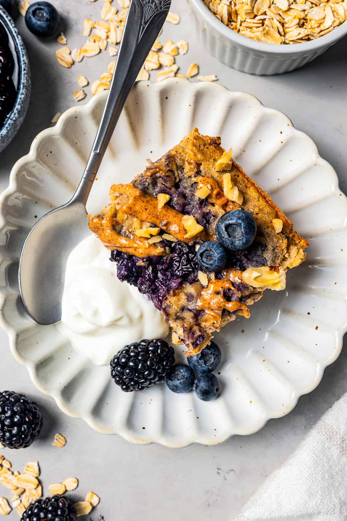 This baked oatmeal is loaded with protein from wholesome oats, almond butter, eggs, and protein powder. It's delicious, swirled with a juicy berry compote. Overhead view of a serving of protein baked oatmeal swirled with berry compote next to yogurt and fresh berries on a plate.