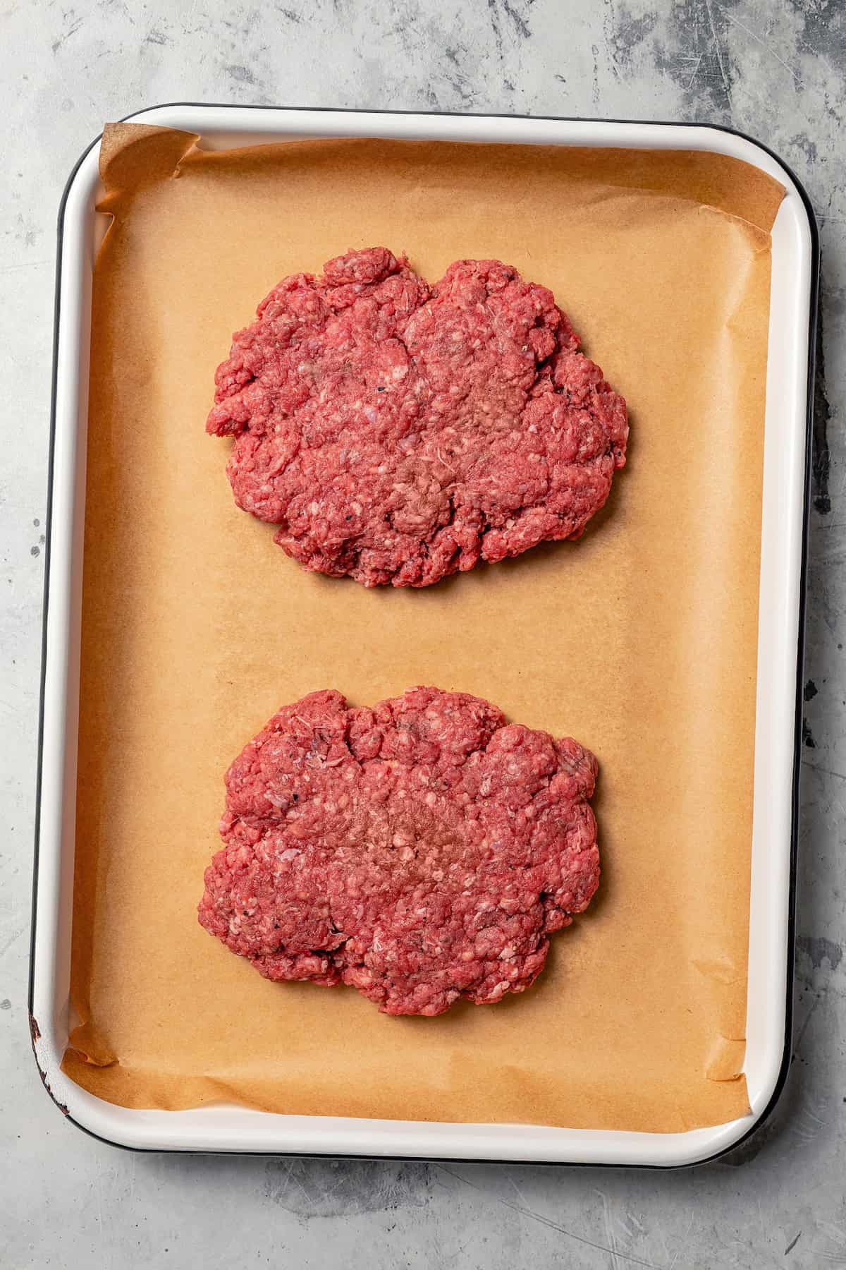 Two ground beef patties pressed flat on a lined baking sheet.
