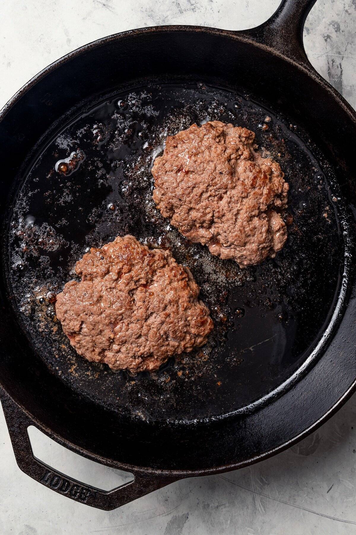 Two ground beef patties browning in a cast iron skillet.