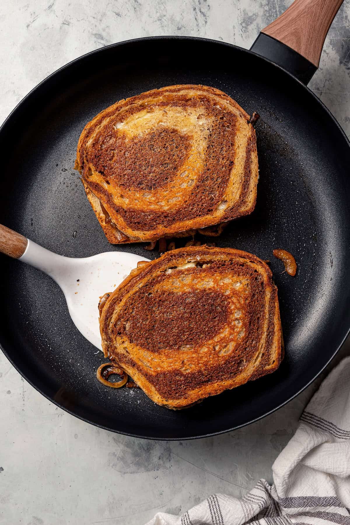 Two patty melt sandwiches toasting in a cast iron skillet, with a spatula tucked underneath one sandwich.