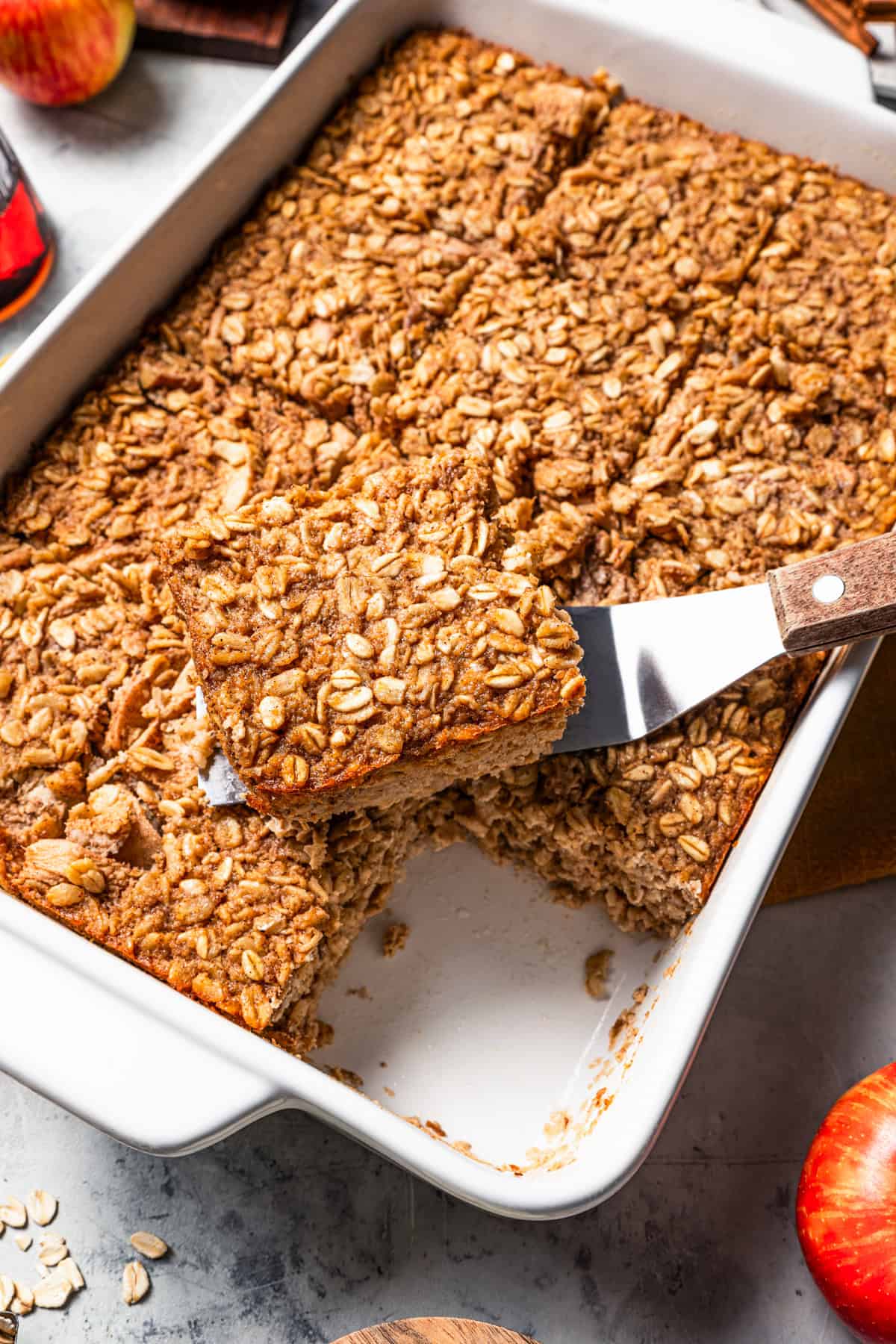 A spatula lifting a slice of apple baked oatmeal over more slices inside a baking dish.