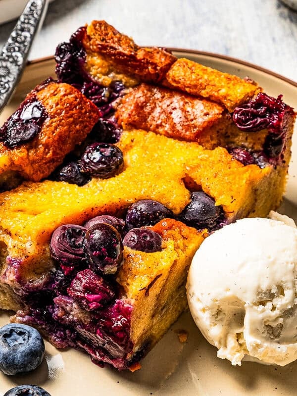 A slice of bread pudding with blueberries served on a plate with a scoop of ice cream.