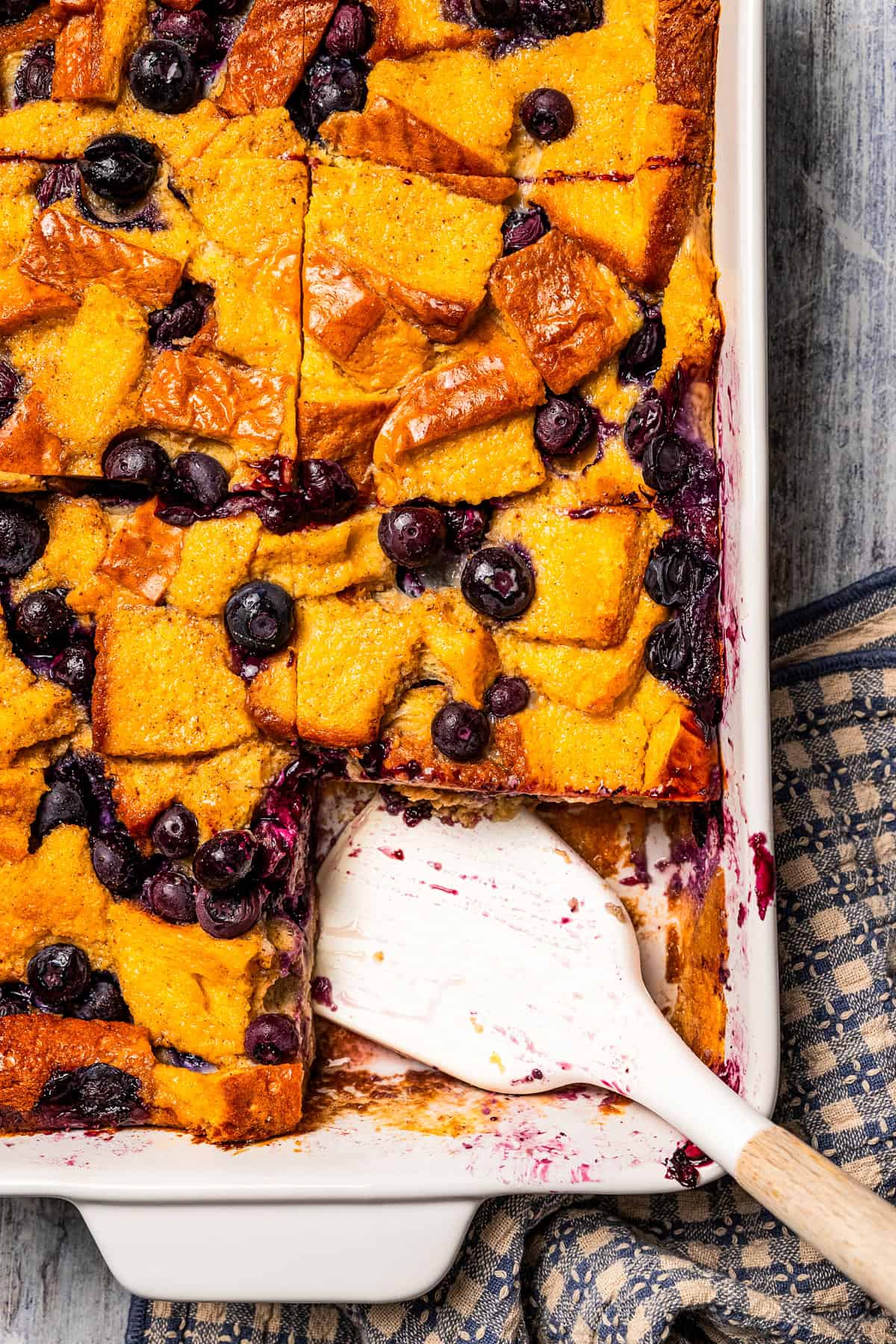 Blueberry bread pudding in a ceramic baking dish with a spatula resting in an open space in the corner.