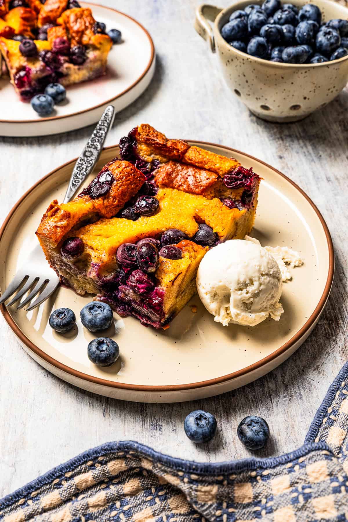 A slice of bread pudding with blueberries served on a plate with a scoop of ice cream.