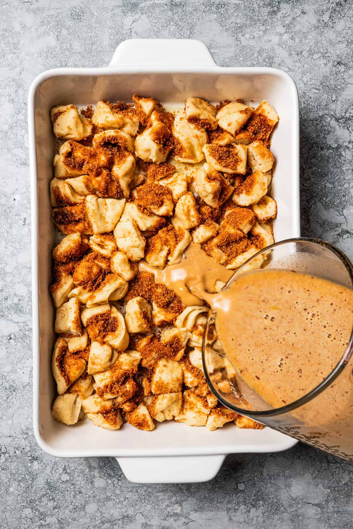 Pouring custard over cinnamon roll pieces in a ceramic baking dish.