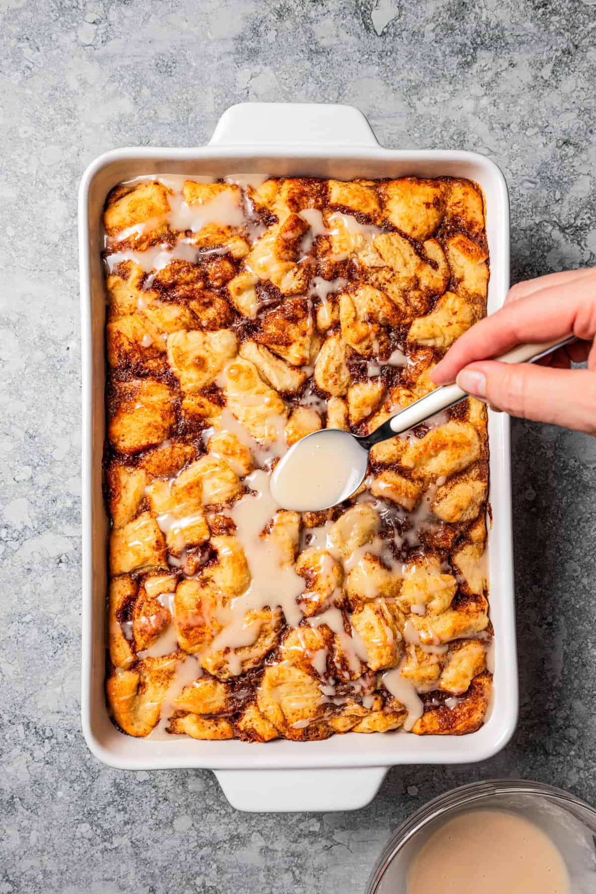 Drizzling icing over a cinnamon roll casserole in a baking dish.