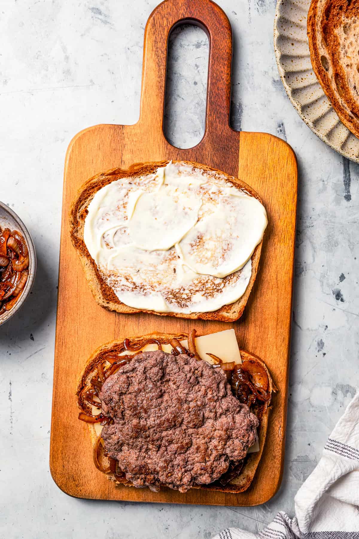 A beef patty on top of cheese and rye next to a second slice of rye covered with mayo on a wooden cutting board.