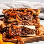 Two halves of a patty melt are stacked on a cutting board next to fries.