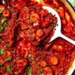 A spoon lifting Swiss steak from tomato gravy in a Dutch oven.