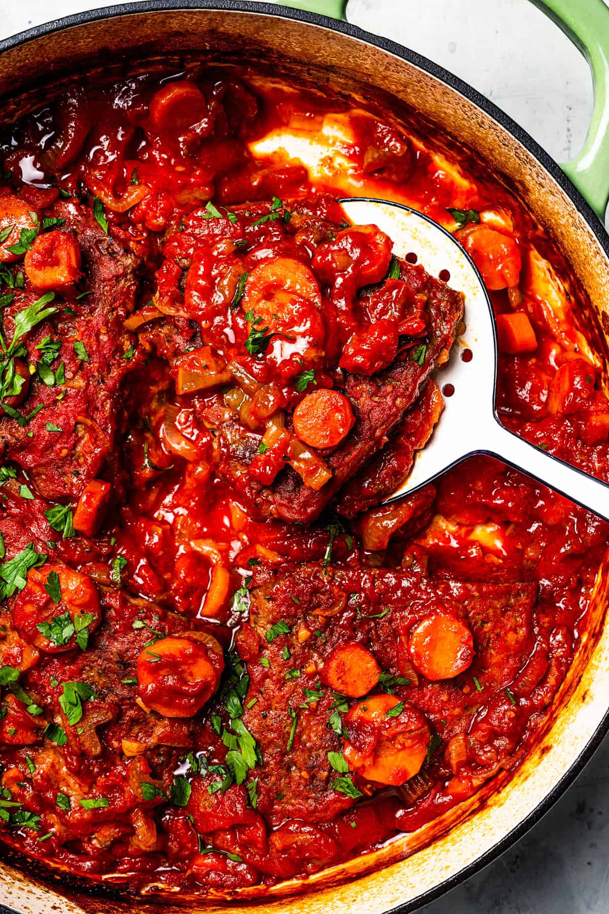 This Swiss steak recipe with gravy is a comforting dinner with juicy beef slow-braised in a rich tomato sauce and served over mashed potatoes. A spoon lifting Swiss steak from tomato gravy in a Dutch oven.