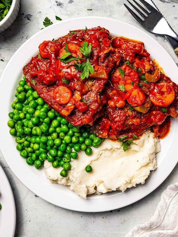 Swiss steak with tomato sauce served with mashed potatoes and green peas on dinner plates.