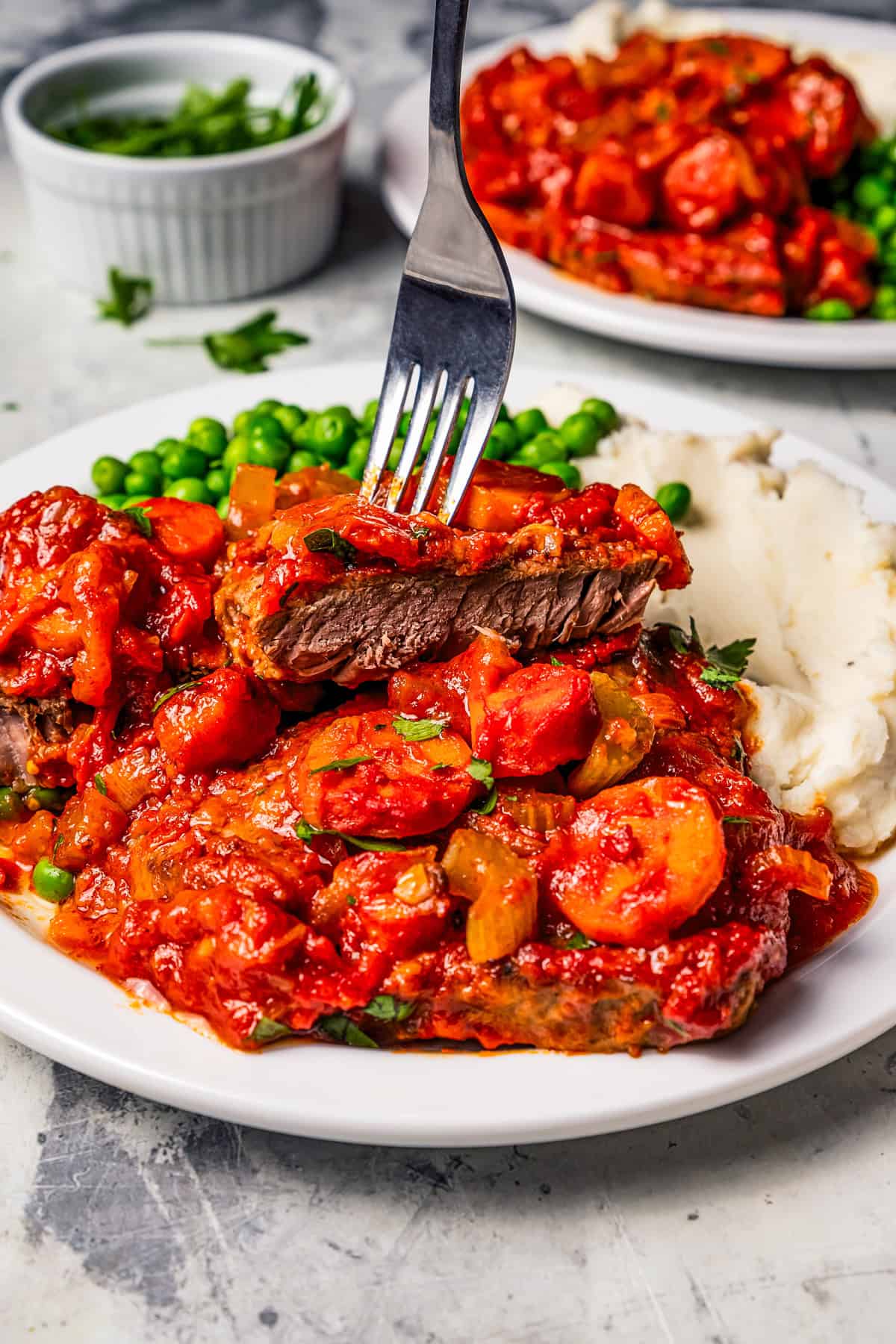 This Swiss steak recipe with gravy is a comforting dinner with juicy beef slow-braised in a rich tomato sauce and served over mashed potatoes. A fork lifting a bite of Swiss steak on a plate.