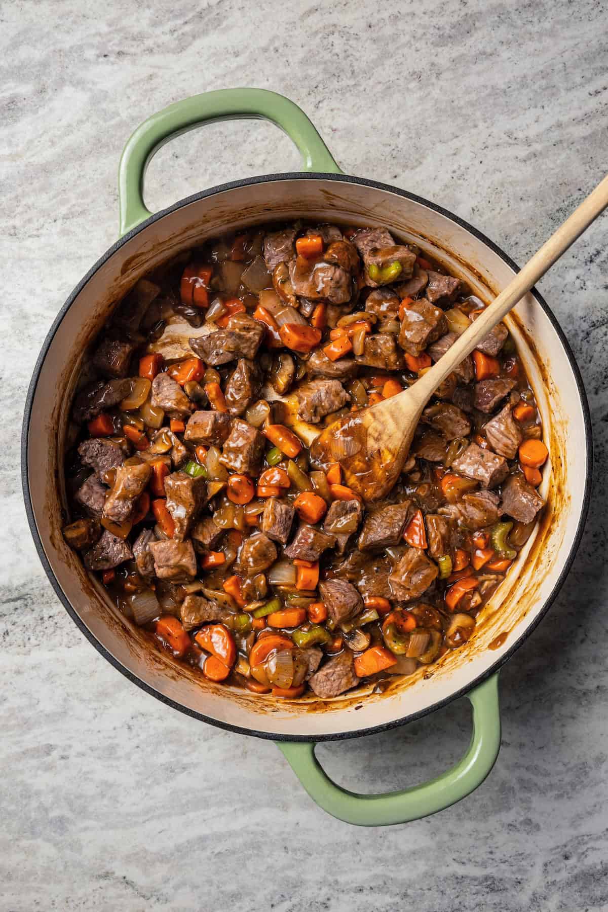This easy beef pot pie recipe has a hearty, savory filling of juicy braised beef and tender veggies baked inside a flaky homemade pie crust. Beef pot pie filling in a skillet with a wooden spoon.