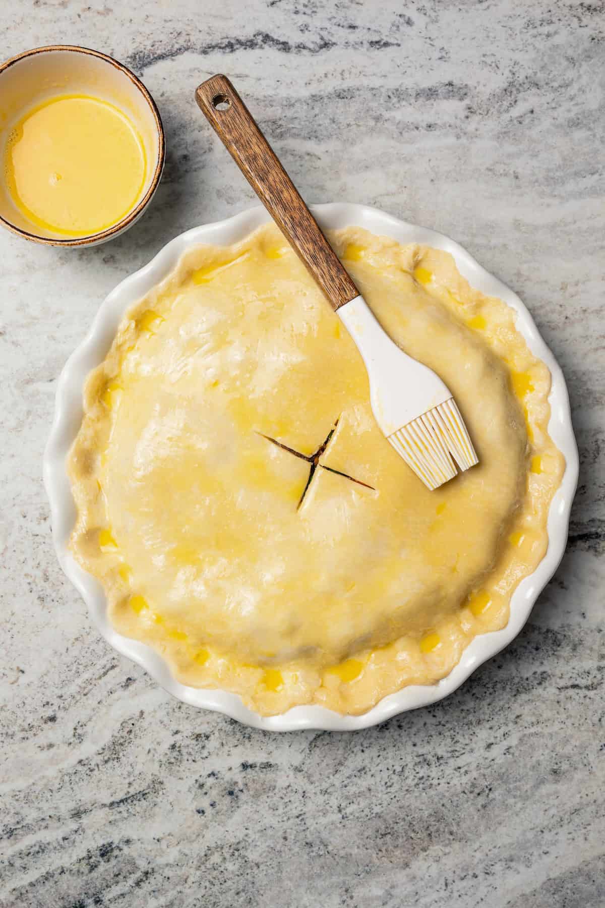 This easy beef pot pie recipe has a hearty, savory filling of juicy braised beef and tender veggies baked inside a flaky homemade pie crust. Assembled beef pot pie brushed with an egg wash in a pie plate.