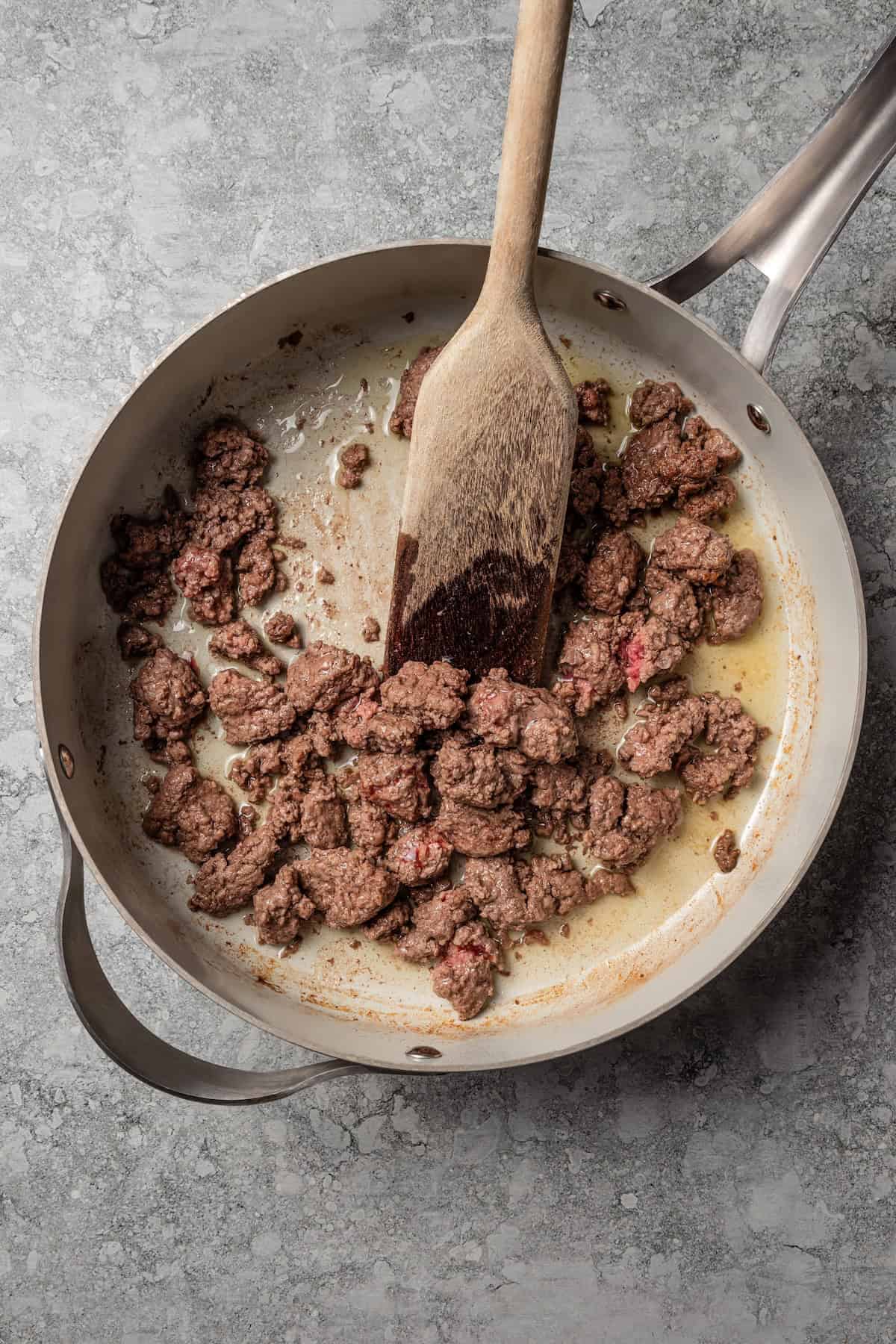 A wooden spoon stirring ground beef while it browns in a skillet.