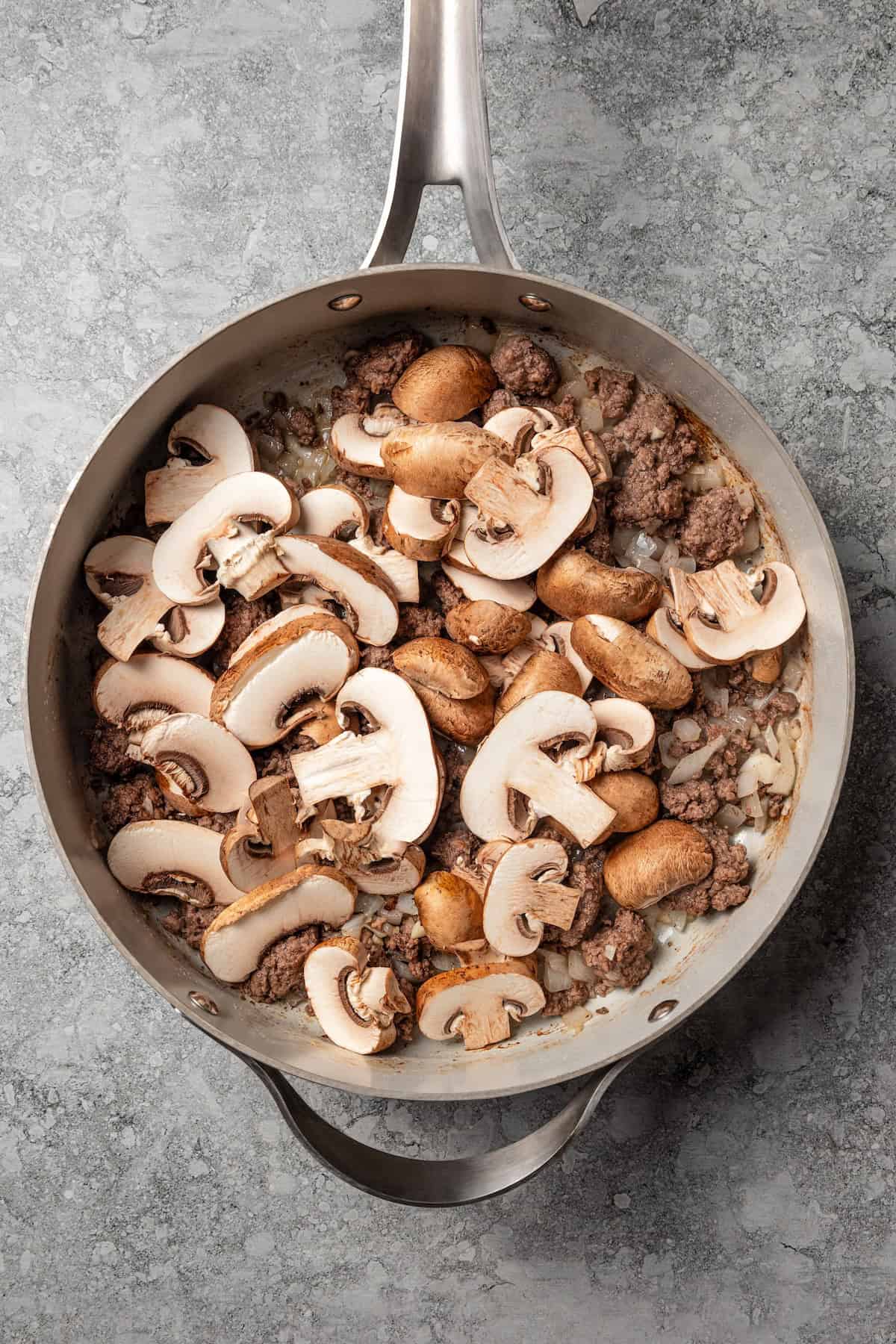 Sliced brown mushrooms added to cooked ground beef and onions a skillet.