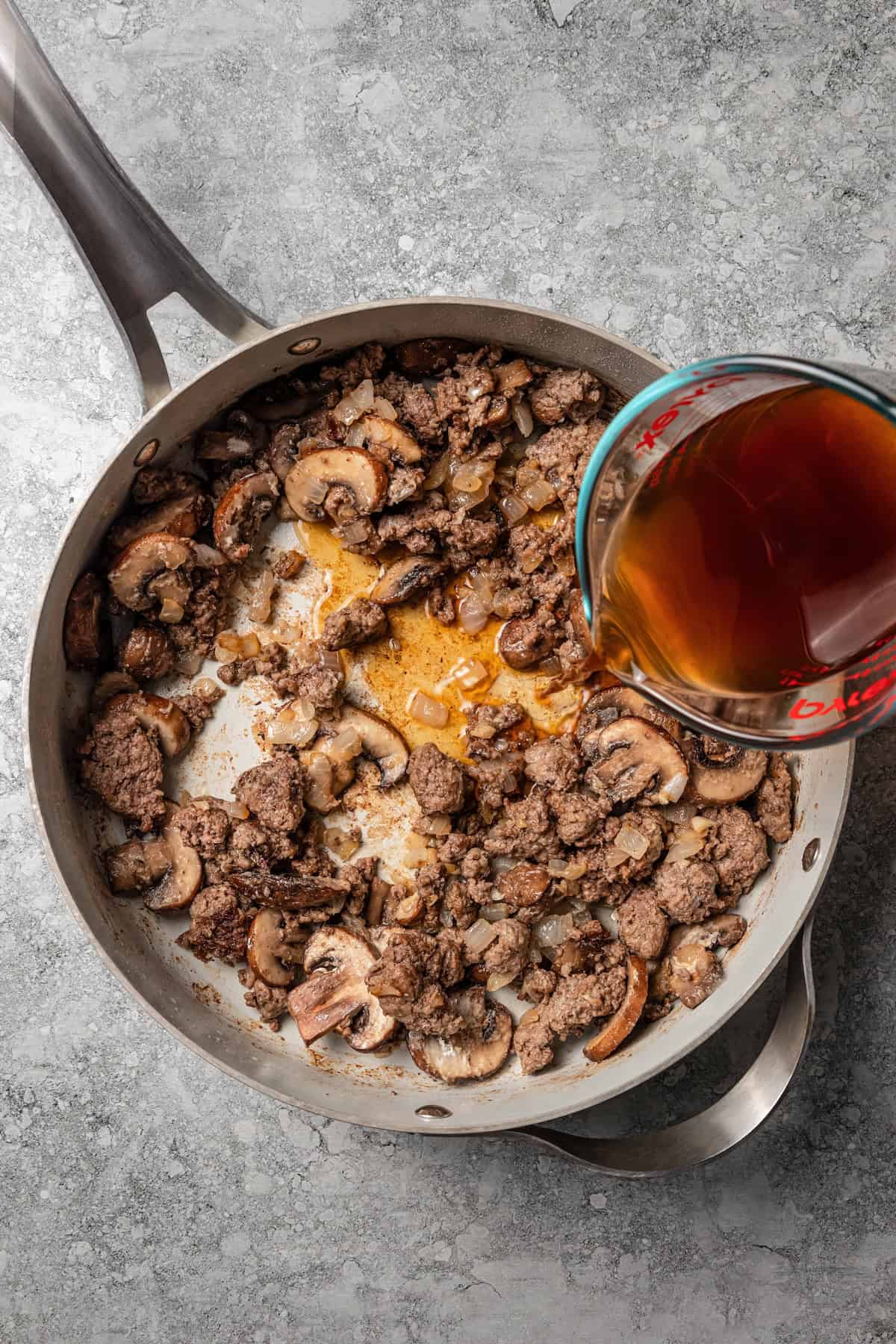 Flour and broth added to sautéed ground beef, mushrooms, and onions in a skillet.