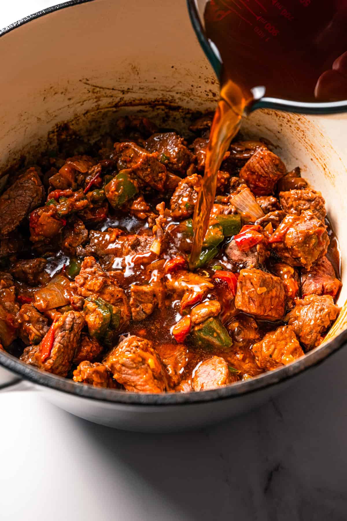 Carne guisada is a flavorful Latin stew with juicy beef slow-cooked in gravy with tomatoes, jalapeños, and spices. Make it at home in one pot! Pouring broth over seared cubed beef and veggies in a large pot.