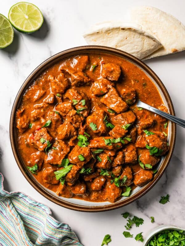 Overhead view of carne guisada in a large bowl with a spoon, next to tortillas and a lime sliced in half.