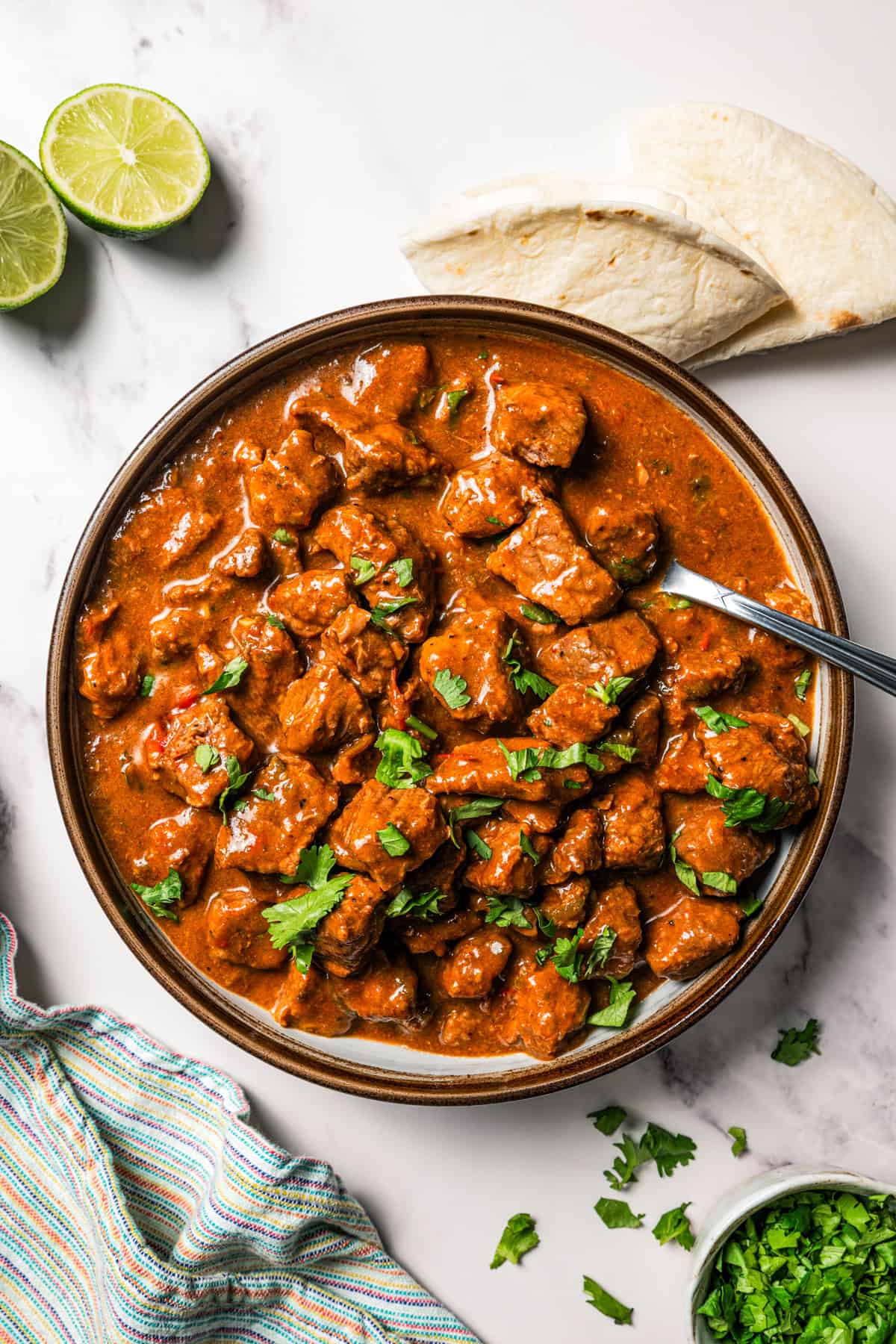 Carne guisada is a flavorful Latin stew with juicy beef slow-cooked in gravy with tomatoes, jalapeños, and spices. Make it at home in one pot! Overhead view of carne guisada in a large bowl with a spoon, next to tortillas and a lime sliced in half.