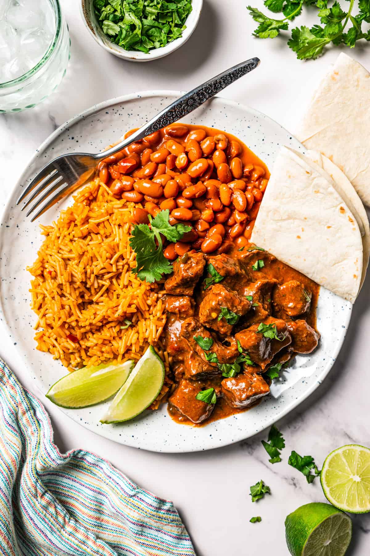 Carne guisada is a flavorful Latin stew with juicy beef slow-cooked in gravy with tomatoes, jalapeños, and spices. Make it at home in one pot! Carne guisada served with Spanish rice, beans, and flour tortillas on a plate.