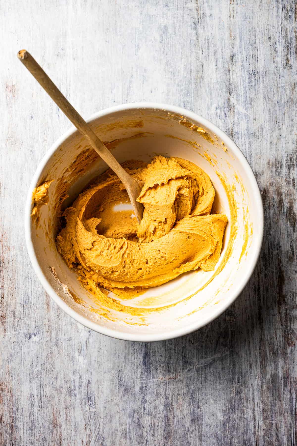 These easy, soft-baked sweet potato cookies are made with sweet potato purée, fall spices, and stuffed with sweet cream cheese in the middle! Sweet potato cookie dough combined in a white bowl with a spoon.