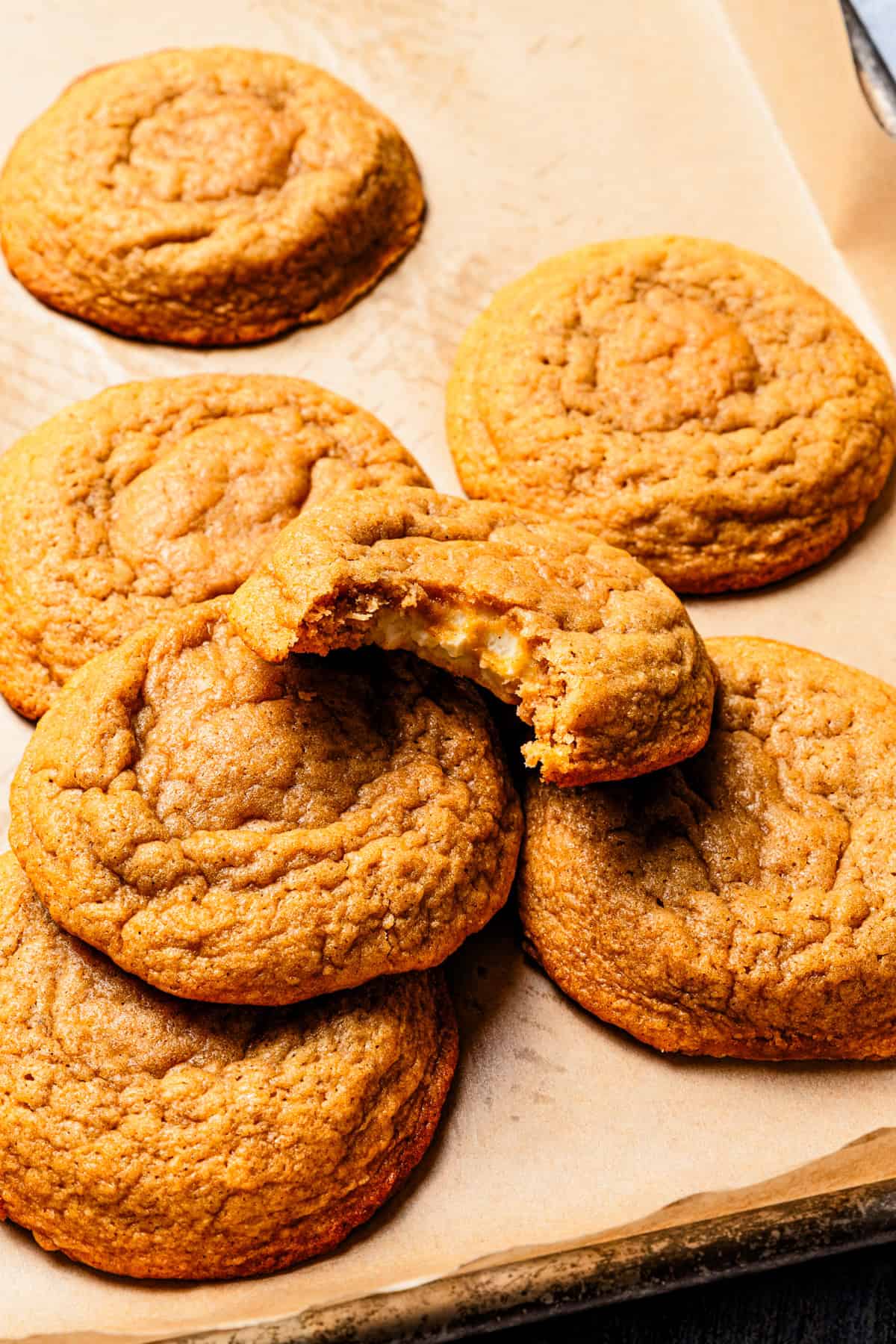 These easy, soft-baked sweet potato cookies are made with sweet potato purée, fall spices, and stuffed with sweet cream cheese in the middle! Sweet potato cookies on a parchment-lined baking sheet, with a bite missing from one cookie.
