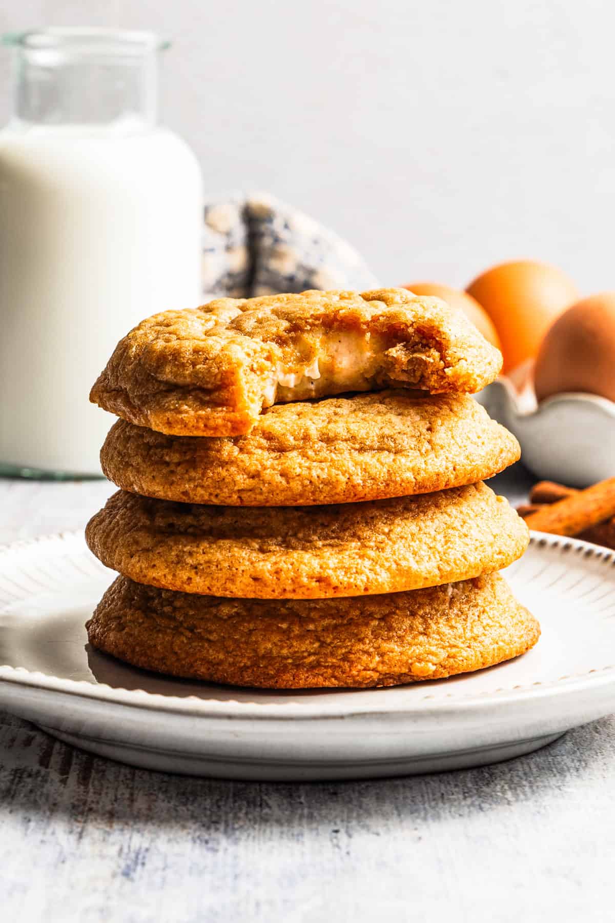 These easy, soft-baked sweet potato cookies are made with sweet potato purée, fall spices, and stuffed with sweet cream cheese in the middle! Sweet potato cookies stacked on a plate with a bite missing from the top cookie, with a glass of milk and carton of eggs in the background.