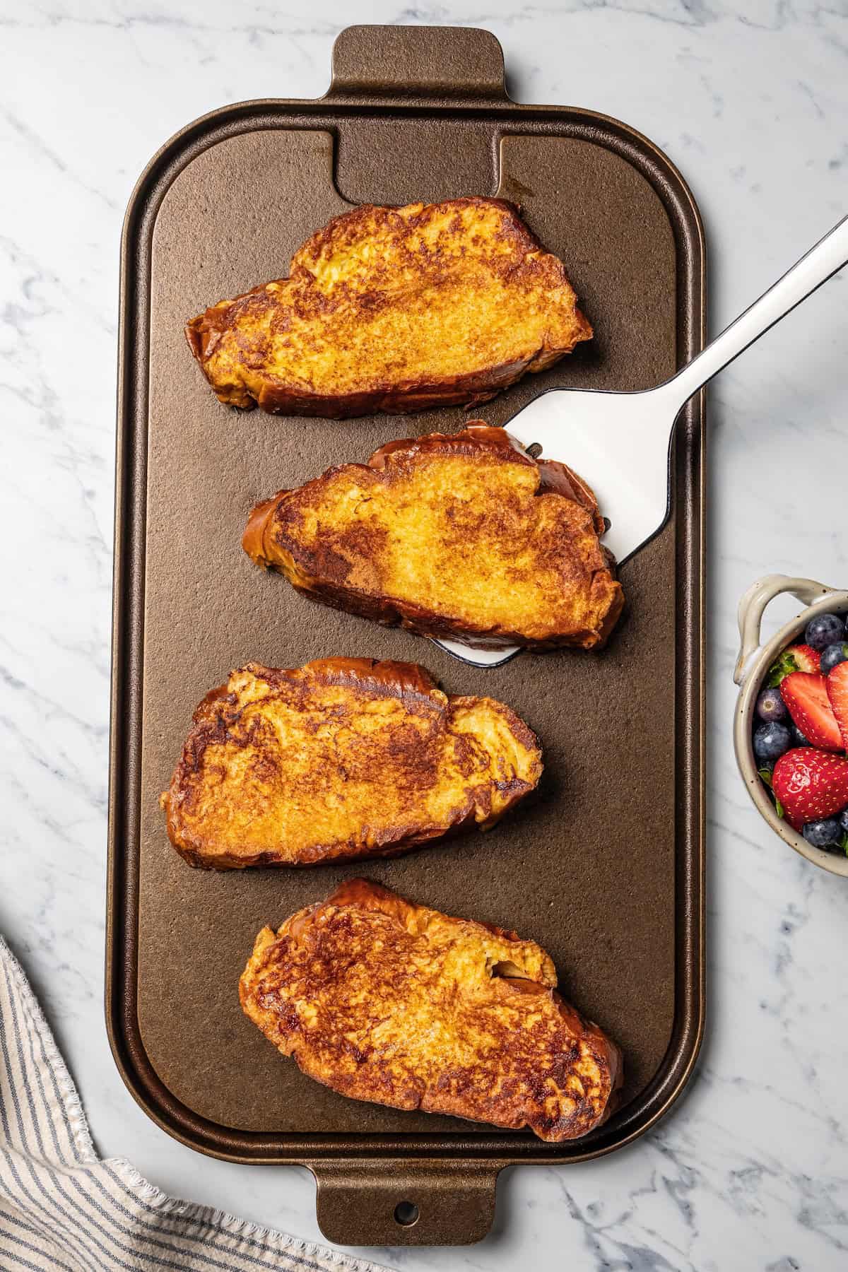 Make thick, fluffy challah bread French toast soaked in an eggy custard with cinnamon, vanilla, and grated nutmeg. The best weekend breakfast! A spatula underneath a slice of challah French toast, next to more slices cooking on a grill pan.