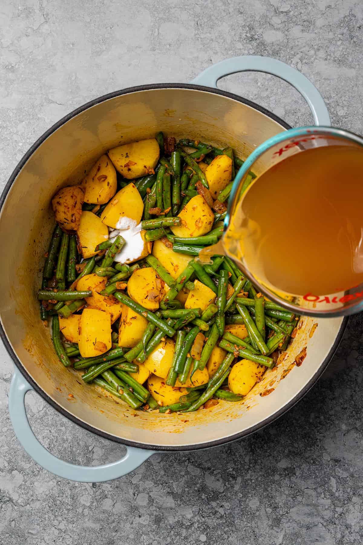 Braised potatoes are slow-cooked with tender green beans and tomatoes in this easy, one-pot Macedonian dish that warms from the inside out. Broth being poured over potatoes and green beans in a pot.