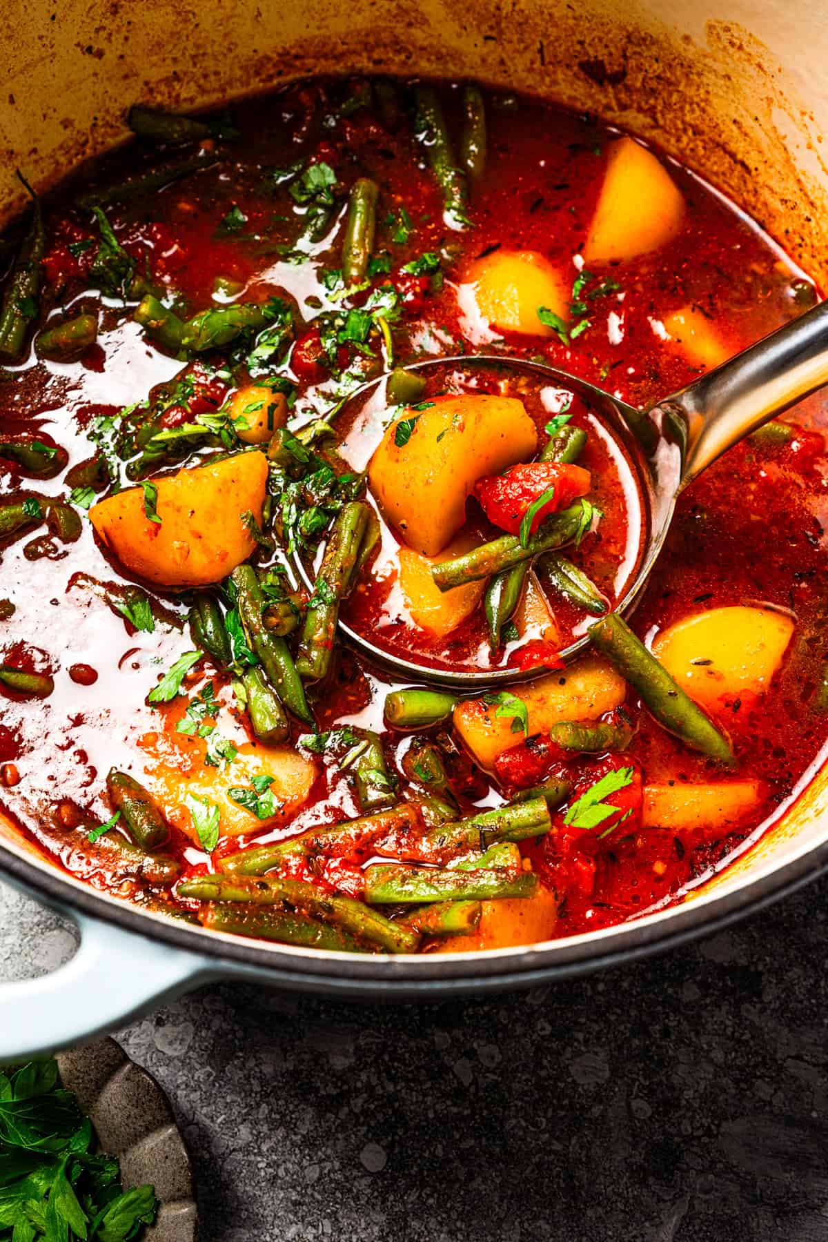 Braised potatoes are slow-cooked with tender green beans and tomatoes in this easy, one-pot Macedonian dish that warms from the inside out. Close up of a ladle lifting Macedonian braised potatoes from a pot.