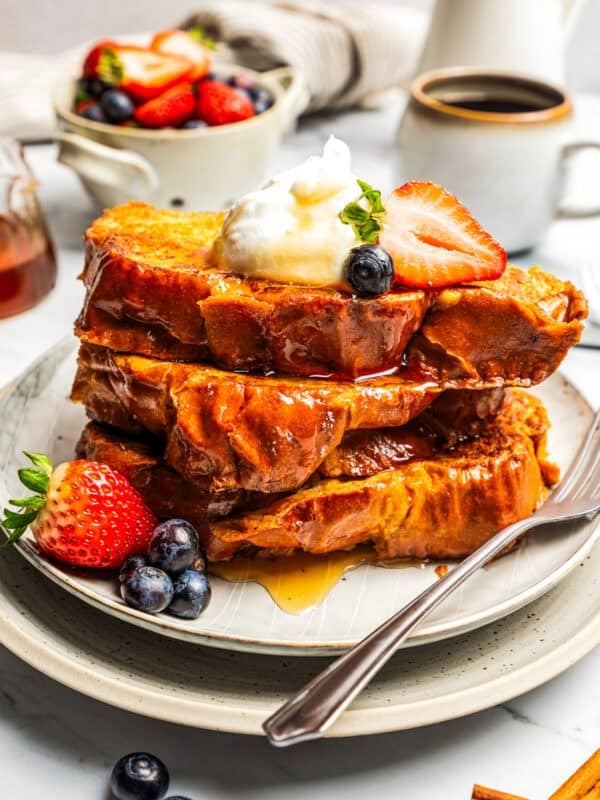 A stack of French toast slices topped with fresh berries, whipped cream, and maple syrup on a plate.