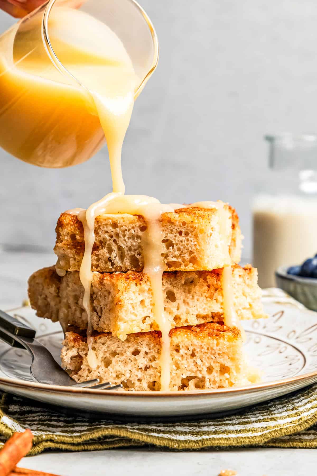 Pouring glaze over a stack of fluffy cinnamon roll sheet pan pancakes on a plate.