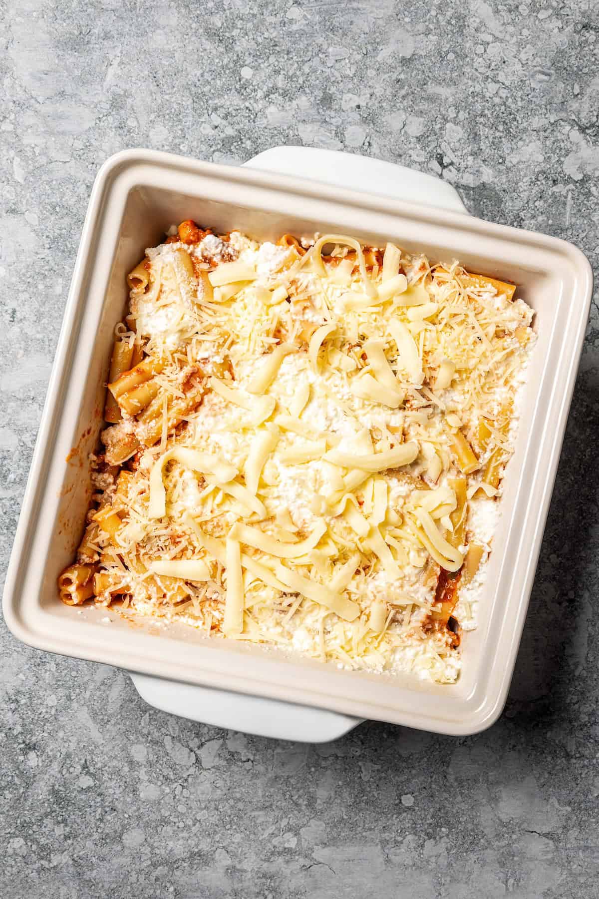 My easy baked ziti recipe is a weeknight-friendly pasta casserole with rich homemade meat sauce and three kinds of melty Italian cheese. Shredded cheese added on top of ziti pasta and sauce in a square baking dish.