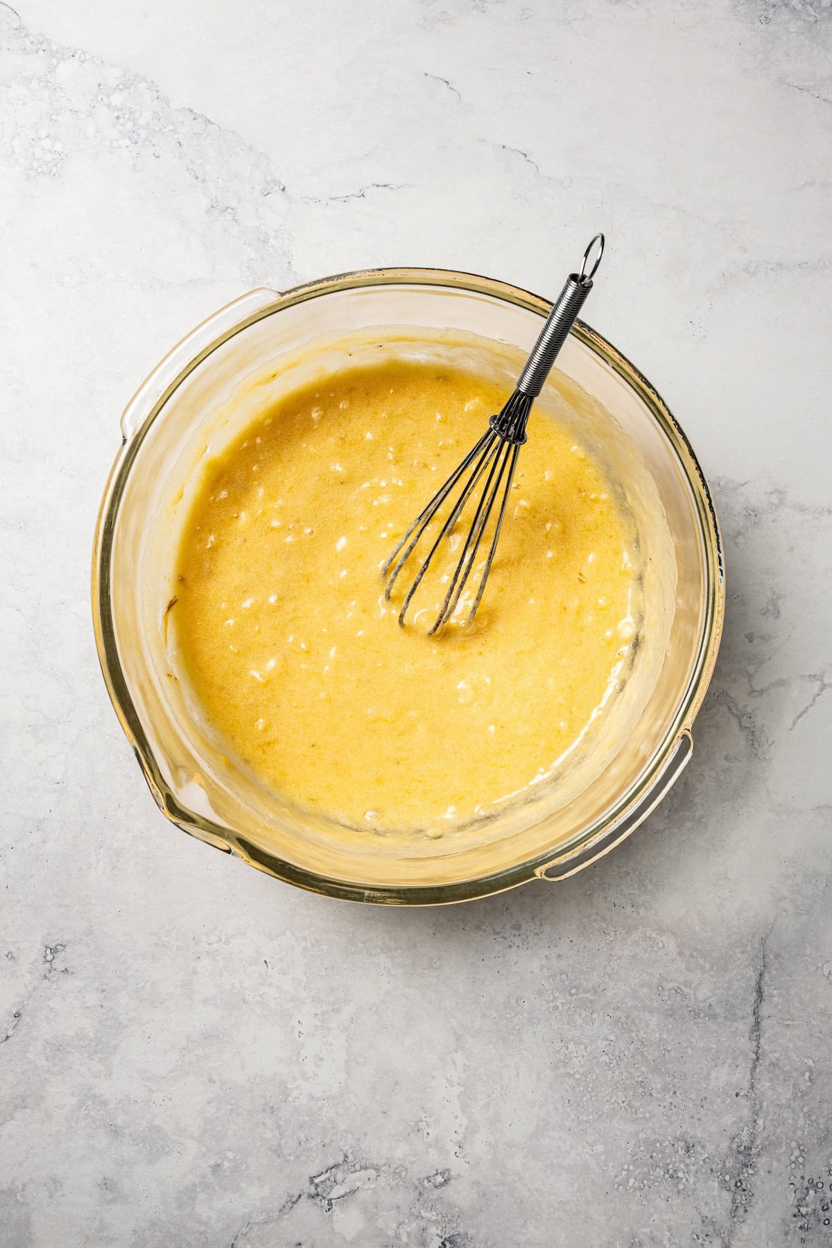 Wet banana muffin batter in a glass mixing bowl with a whisk.